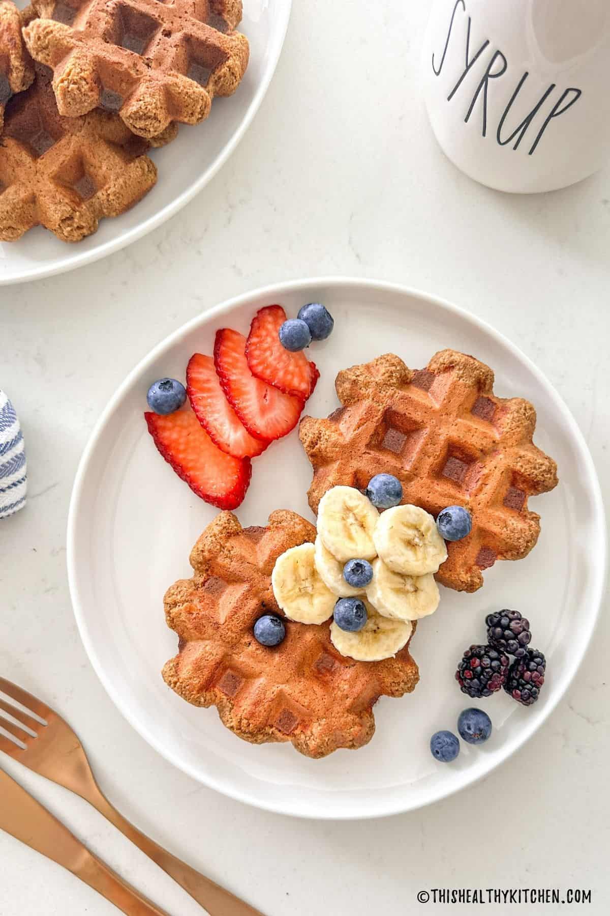 Two waffles on white plate with sliced bananas, strawberries, blackberries and blueberries on top.