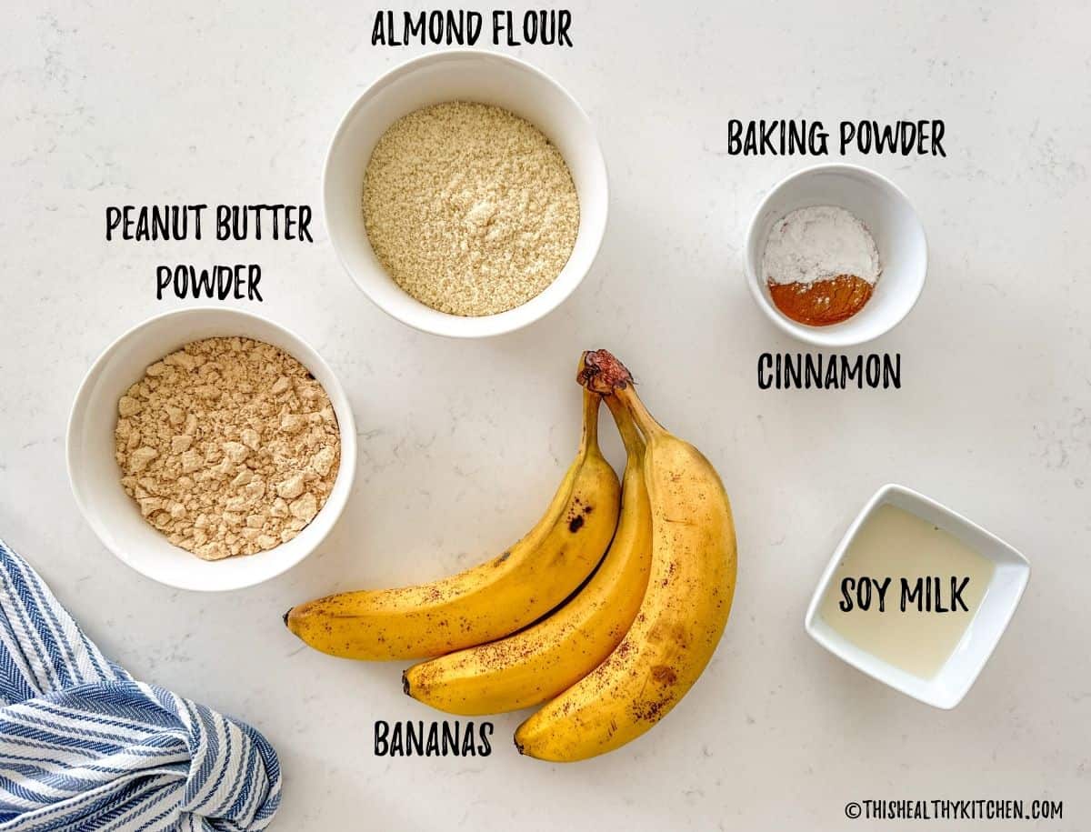Almond flour, peanut butter powder, soy milk, baking powder, cinnamon, and bananas on countertop.