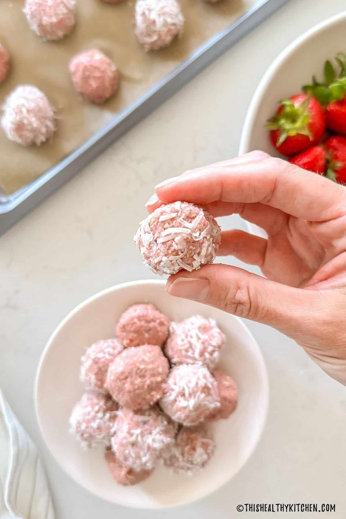 Plate of strawberry energy balls with 1 ball being held above it.