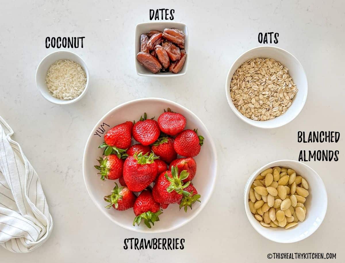 Bowls of fresh strawberries, dates, oats, slivered almonds and coconut shreds on countertop.