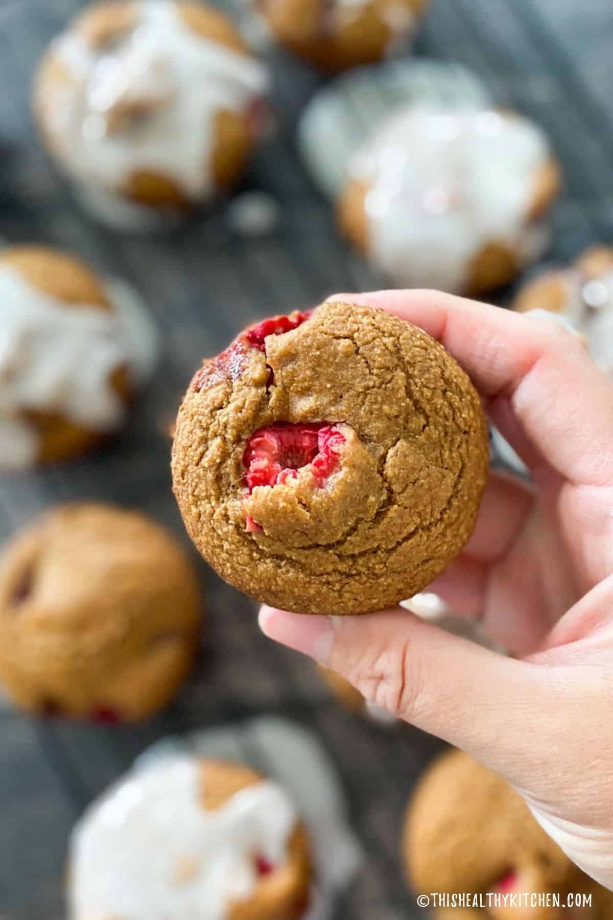 Hand holding up a raspberry muffin with more muffins below it on wire rack.