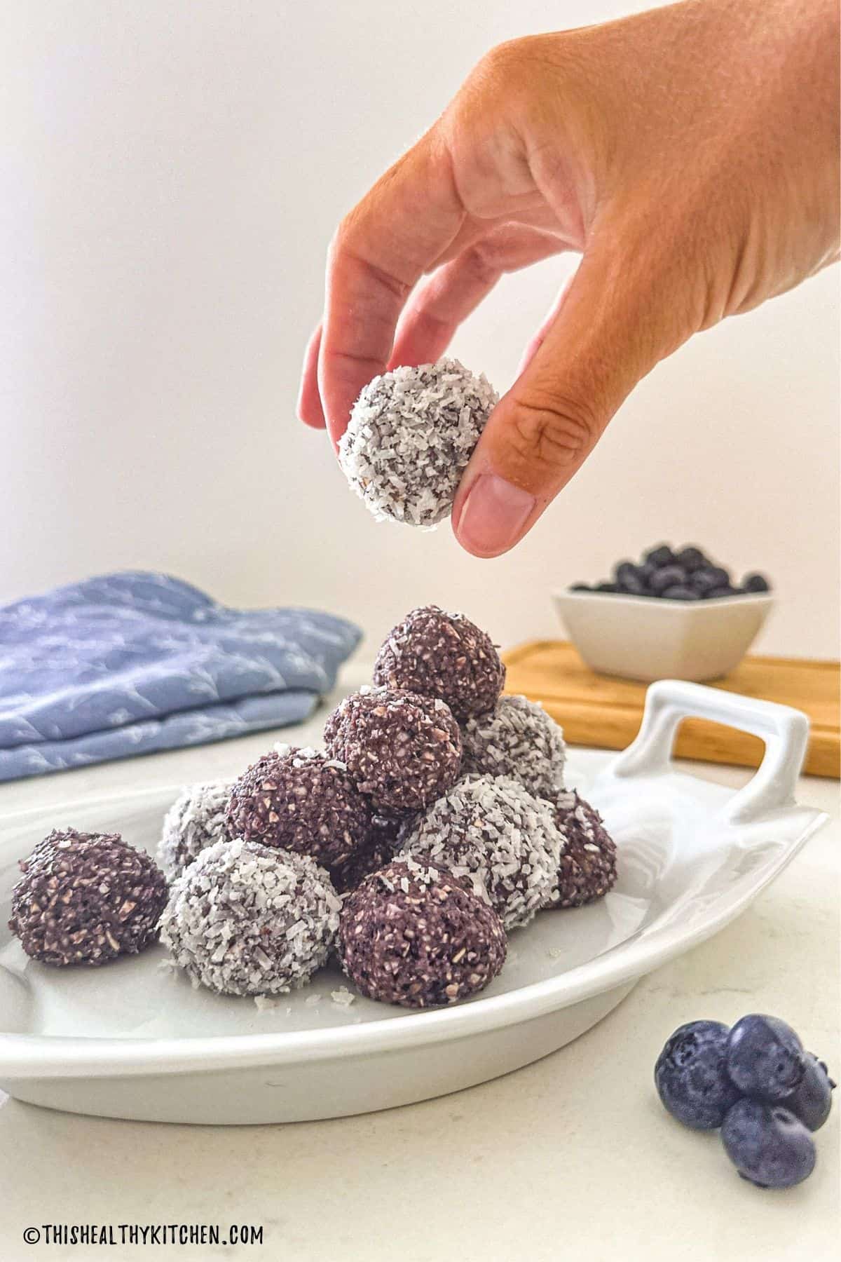 Hand lifting a blueberry energy ball from white platter.