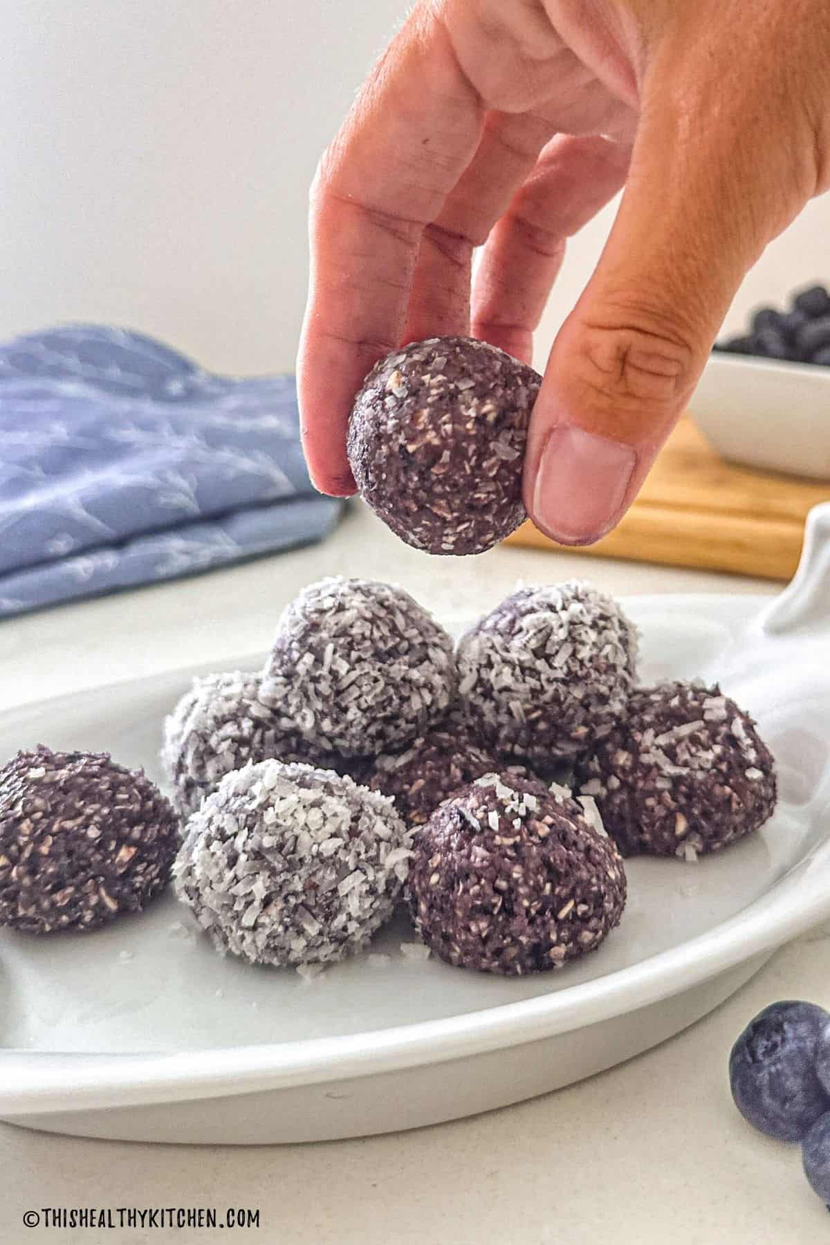 Hand lifting a blueberry energy ball from white platter.