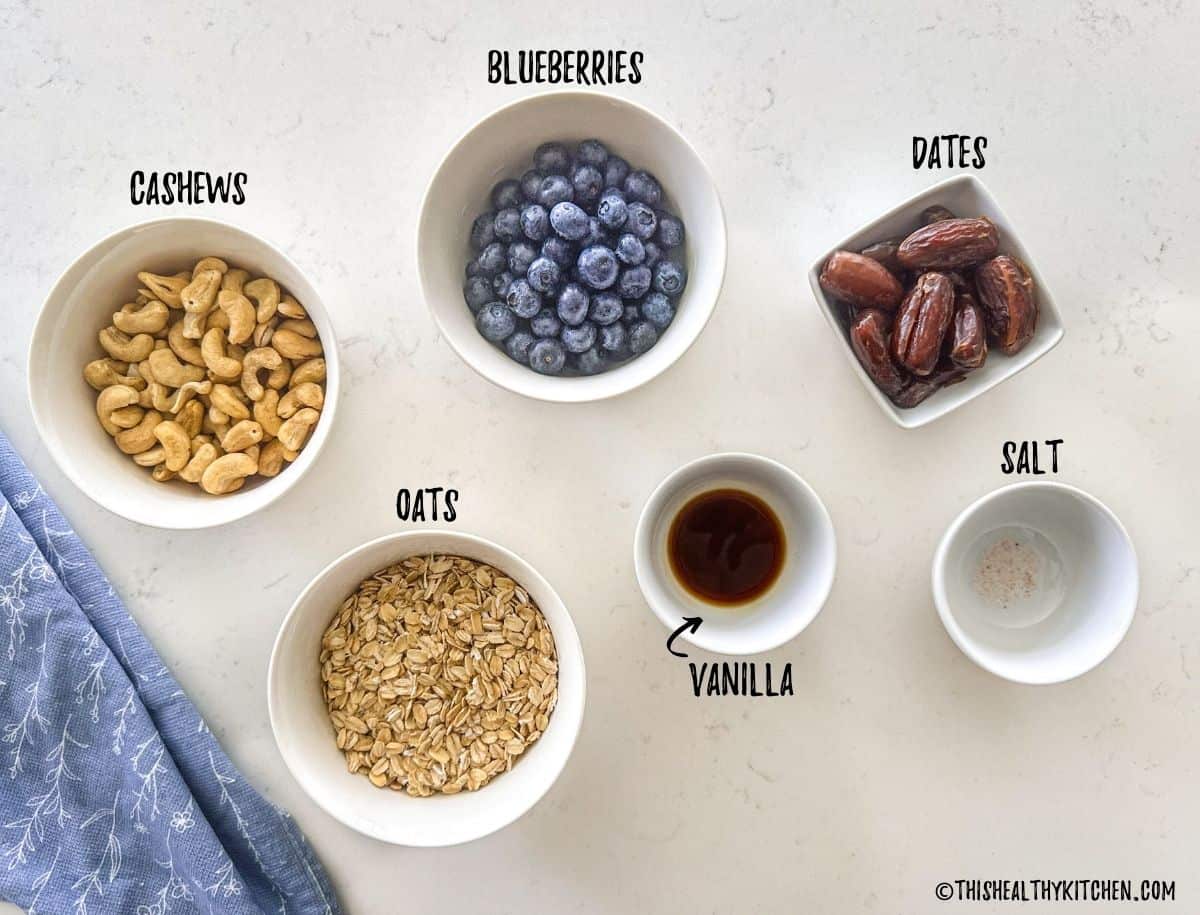 Bowls of cashews, oats, blueberries, dates, vanilla, and salt on kitchen counter.