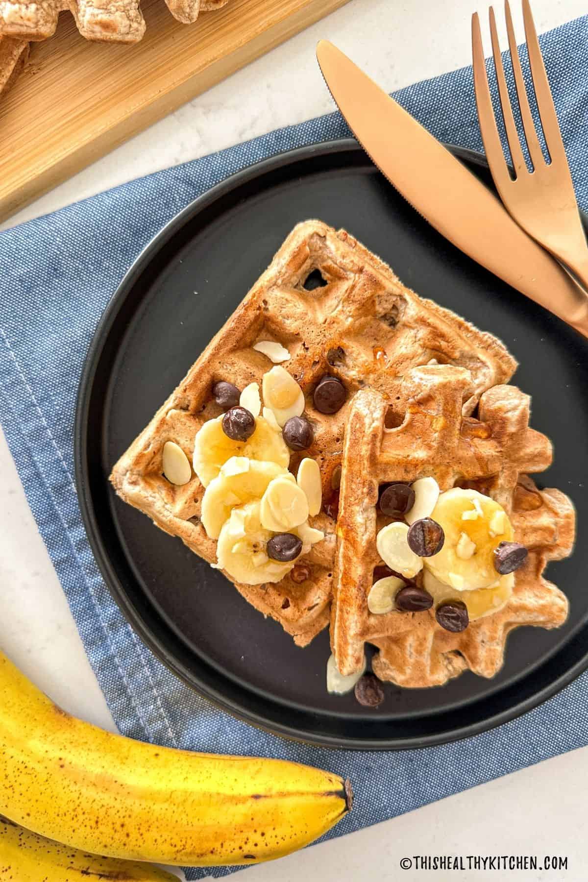 Two banana waffles in black plate with chocolate chips and fork and knife beside it.