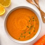 Bowl of carrot and lentil soup with spoons and halved lemons beside it.