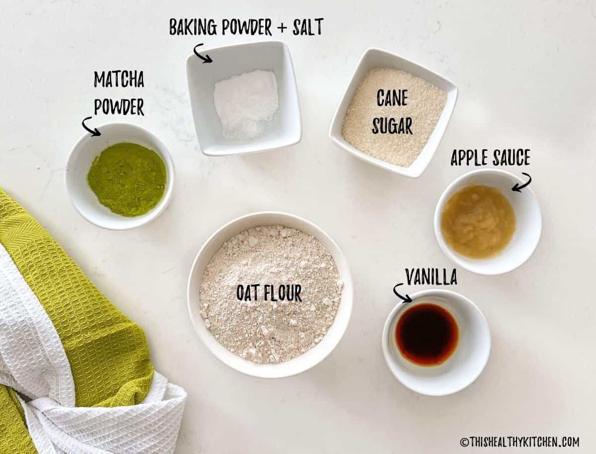 Bowls of oat flour, matcha powder, vanilla, sugar, apple sauce, and baking powder on countertop.