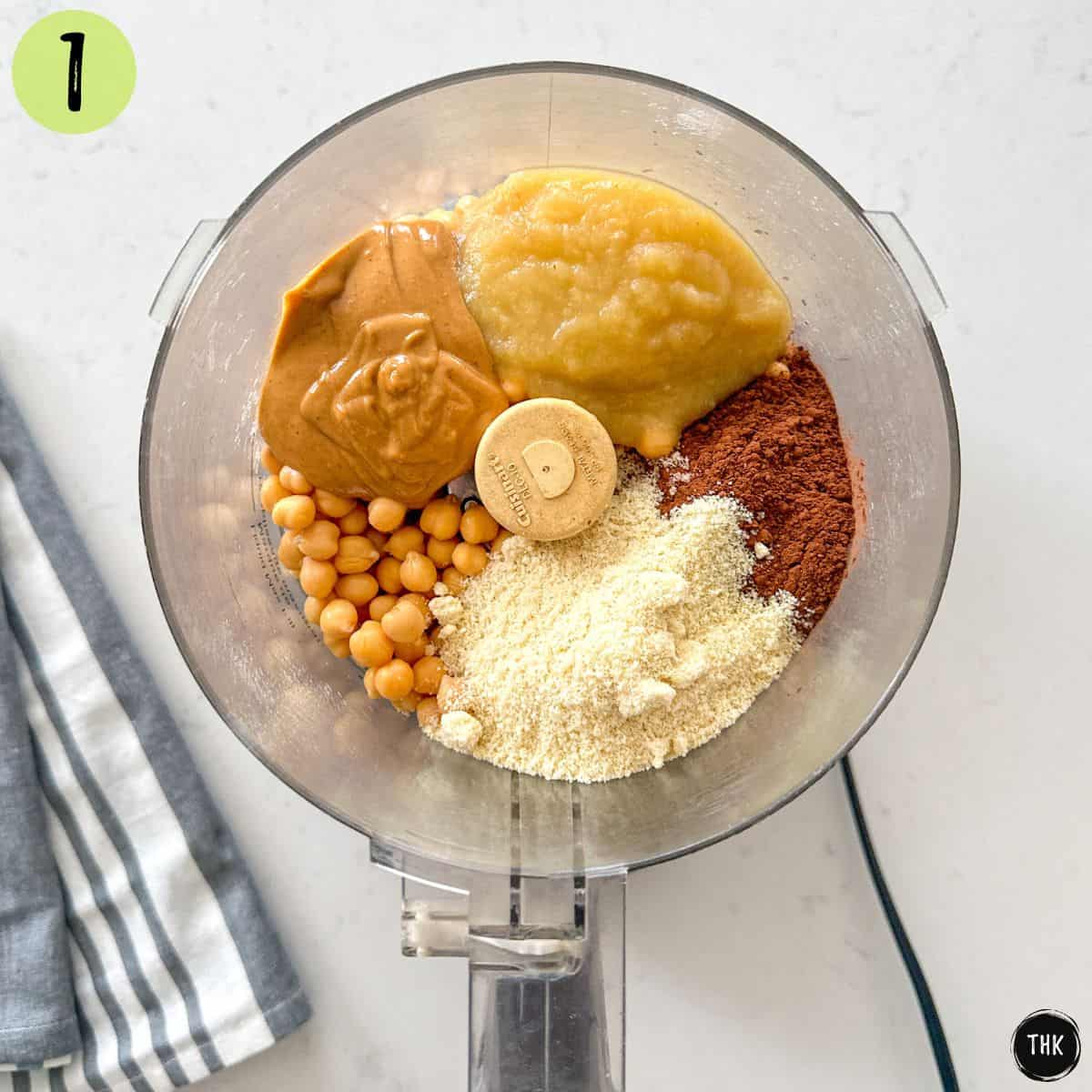 Food processor with flour, cocoa, chickpeas, almond butter, and applesauce inside.