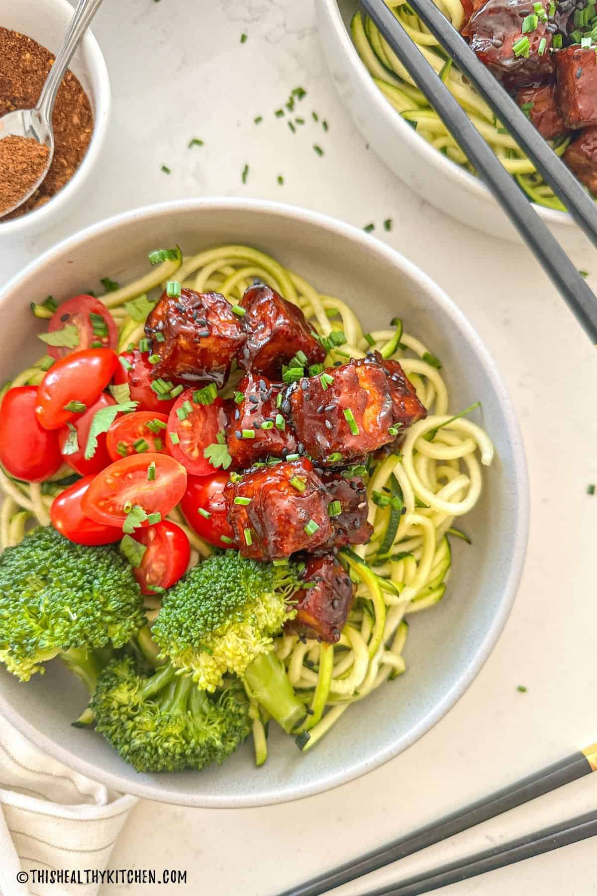 Bowls of zucchini noodles with broccoli, grape tomatoes and spicy tofu on top.