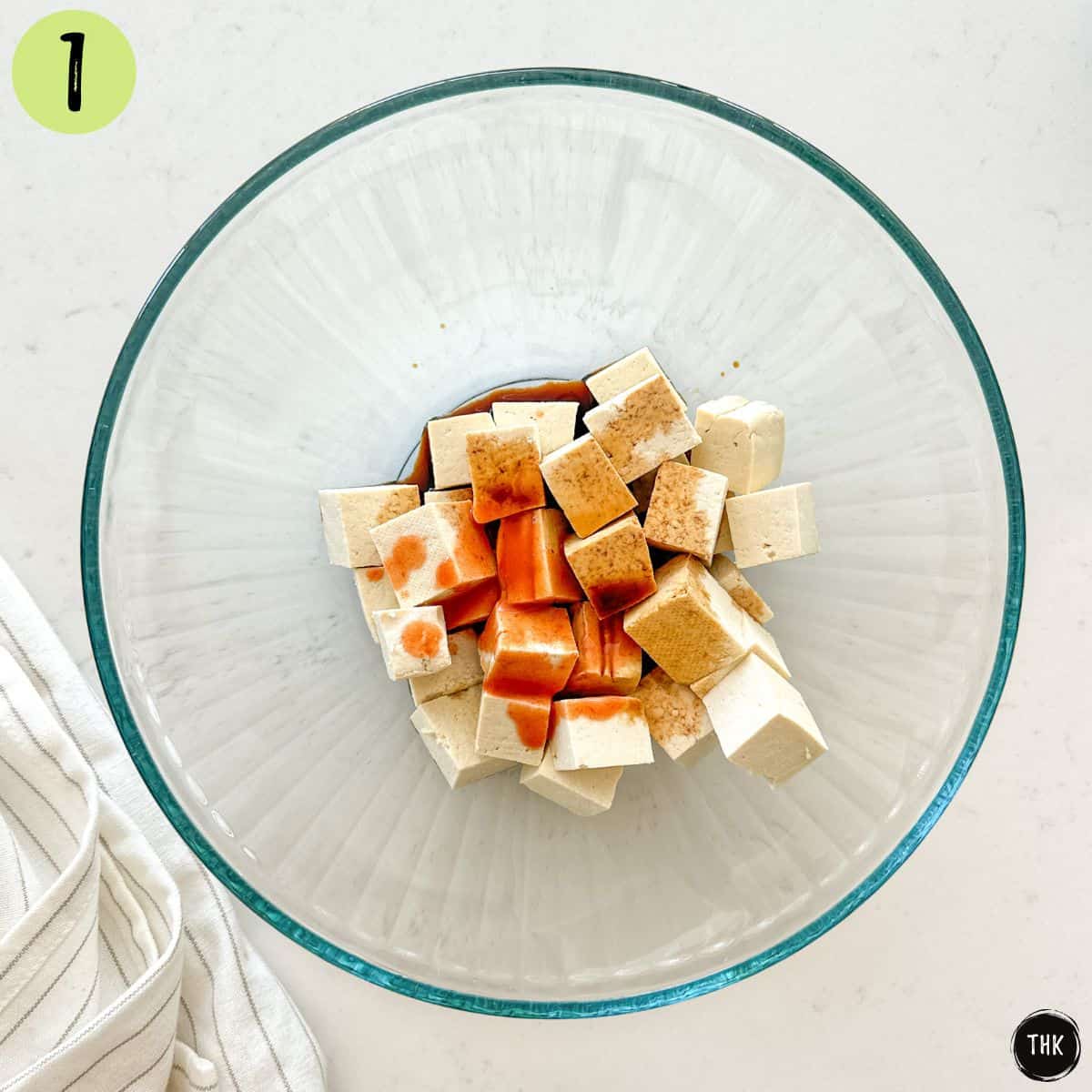 Glass bowl with cubed tofu and soy sauce and hot sauce inside.