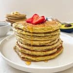 Stack of avocado pancakes on white plate with sliced strawberries on top.