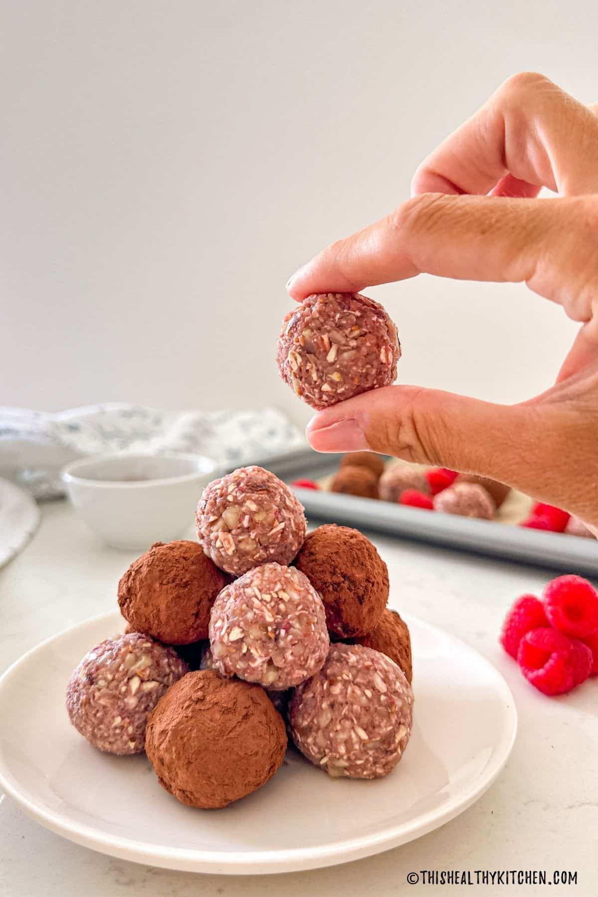 Hand holding up a raspberry bliss ball with more of them on plate below.