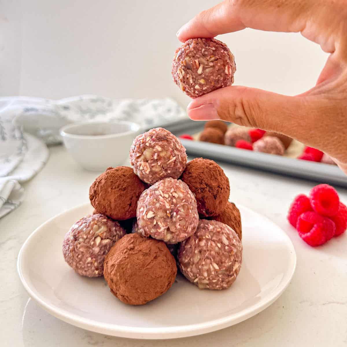 Hand holding up a raspberry bliss ball with more of them on plate below.