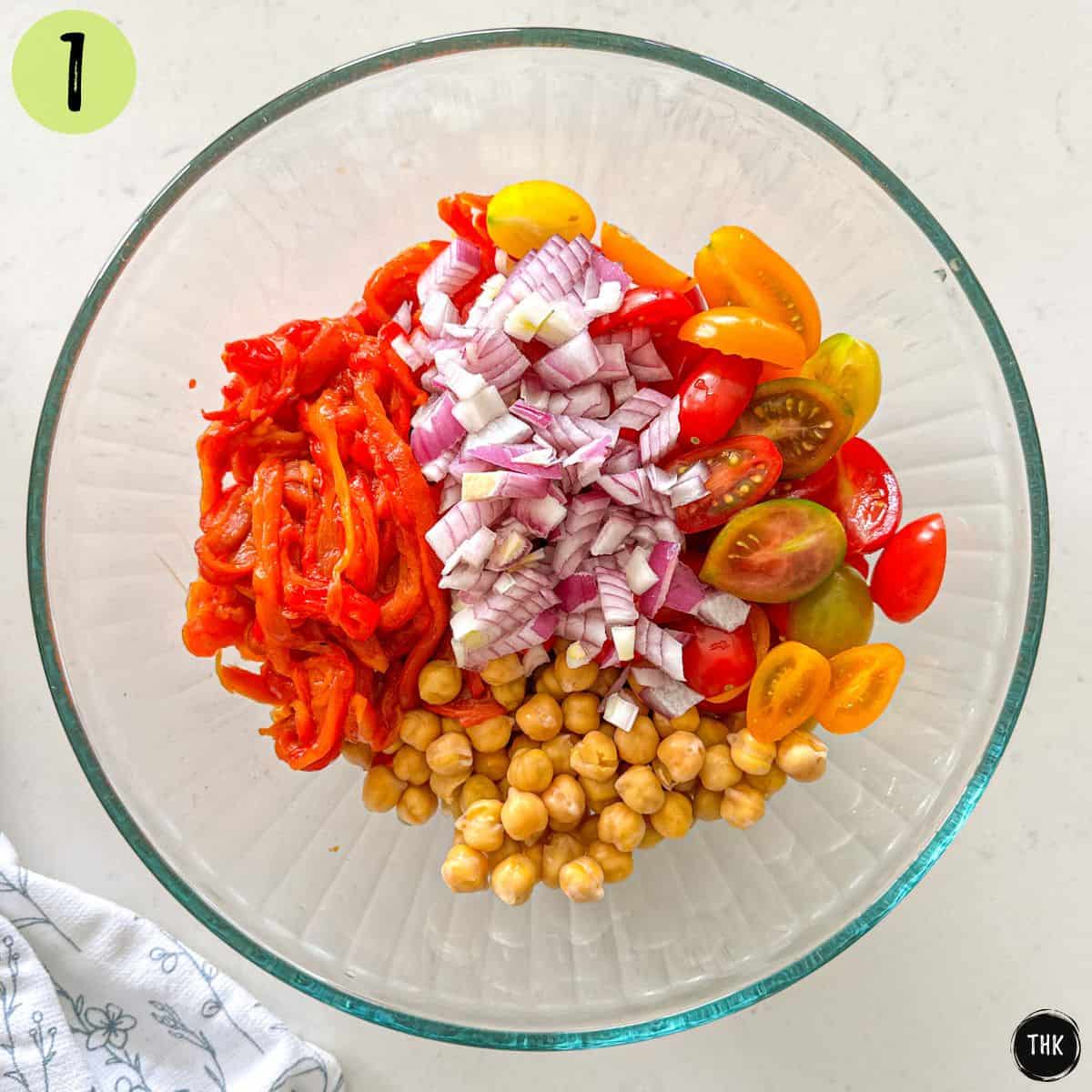 Large glass bowl with roasted red pepper strips, chickpeas, halved cherry tomatoes and diced red onion.