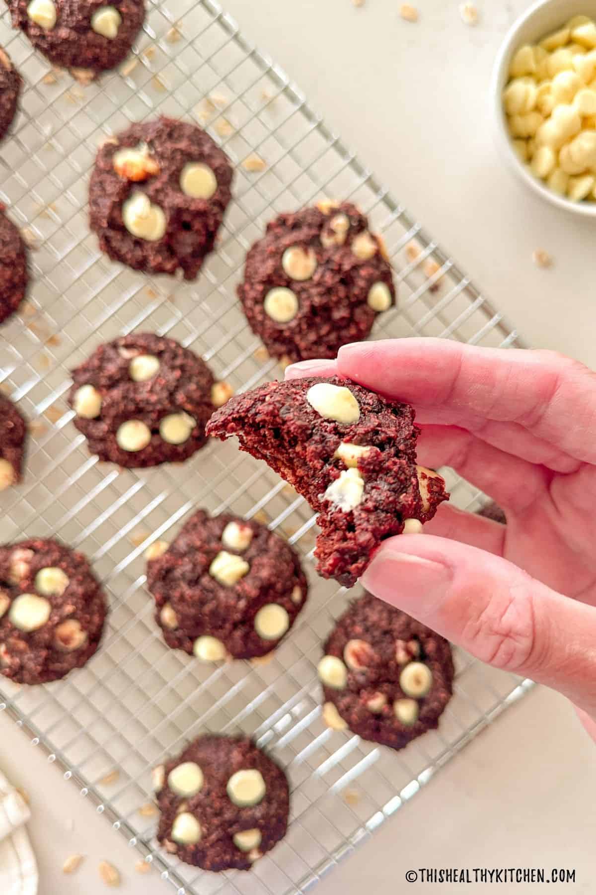 Hand holding up a beet cookie with bite taken out with more cookies below it on cooling rack.