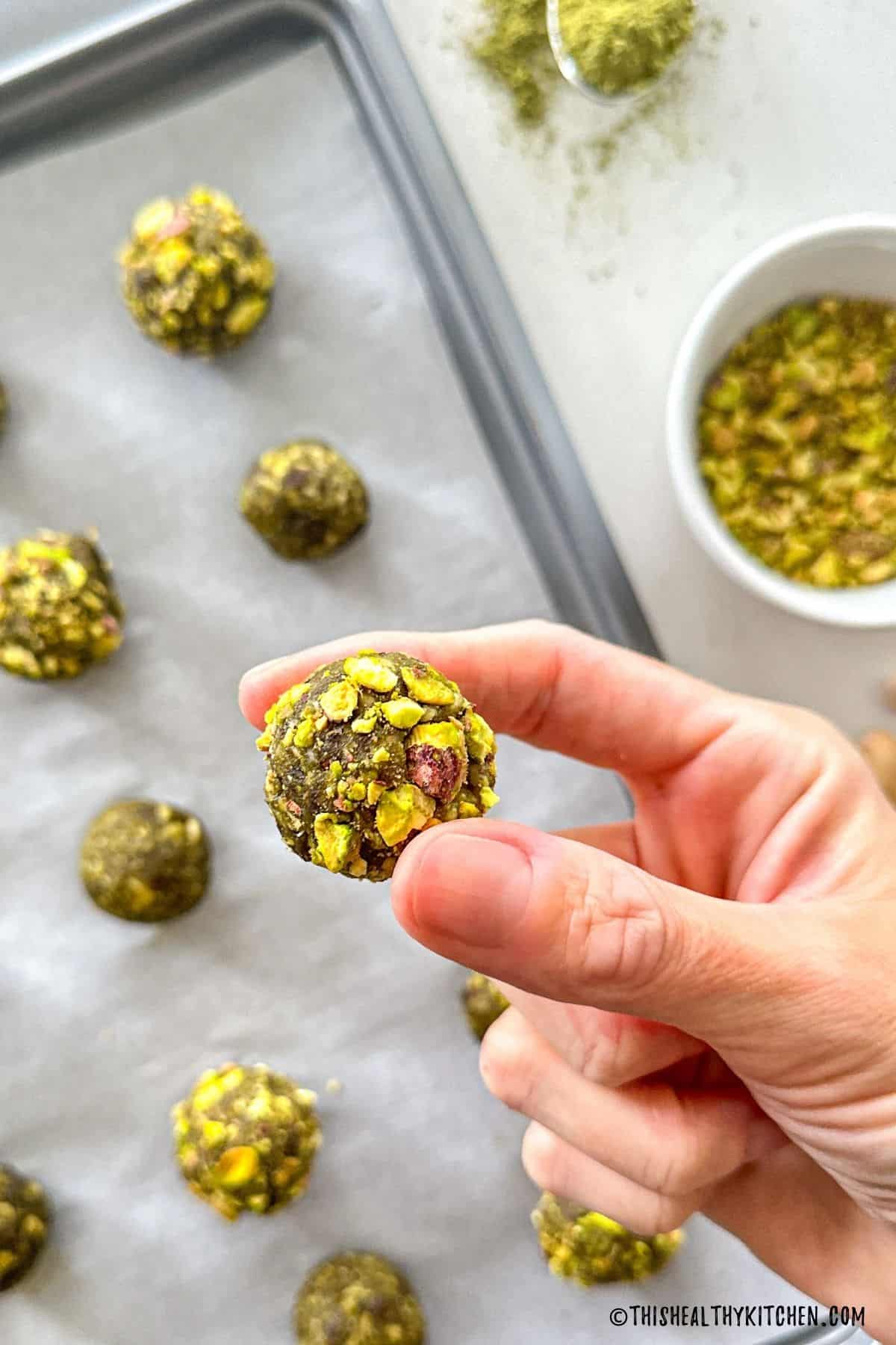 Hand holding up 1 matcha energy ball with tray of more balls below.