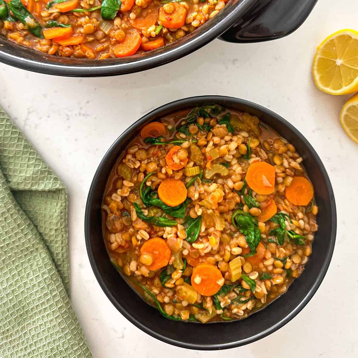 Pot of barley and lentil soup with vegetables and bowl of soup beside it.